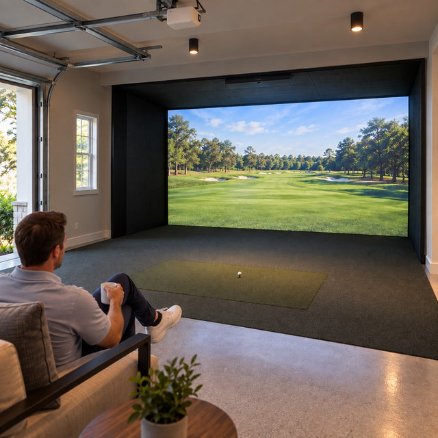 Converted garage golf simulator with a built-in impact screen filling the end wall, golfer seated watching a tree-lined fairway