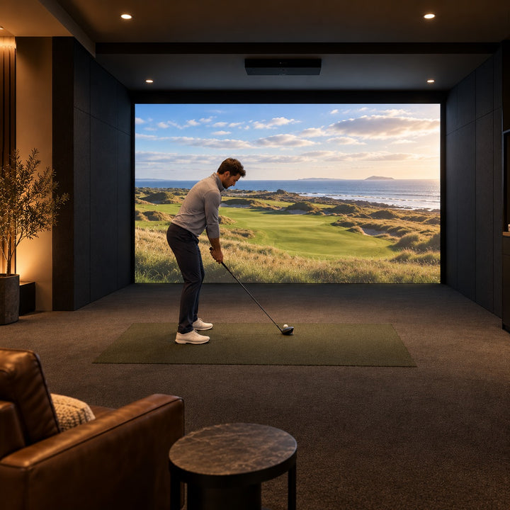 Golfer at address in a dark finished basement golf simulator, built-in impact screen spanning the full wall with a coastal links hole projected